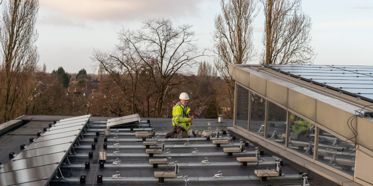 A man wearing a yellow high-vis jacket works on a roof covered with solar panels.