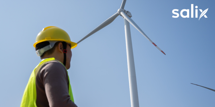 Engineer in safety equipment standing near a wind turbine at a renewable energy installation.
