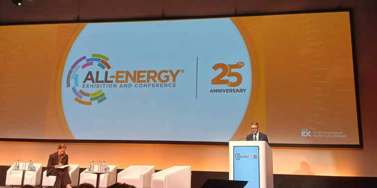 A man addresses a conference from a lecturn in front of an orange background.