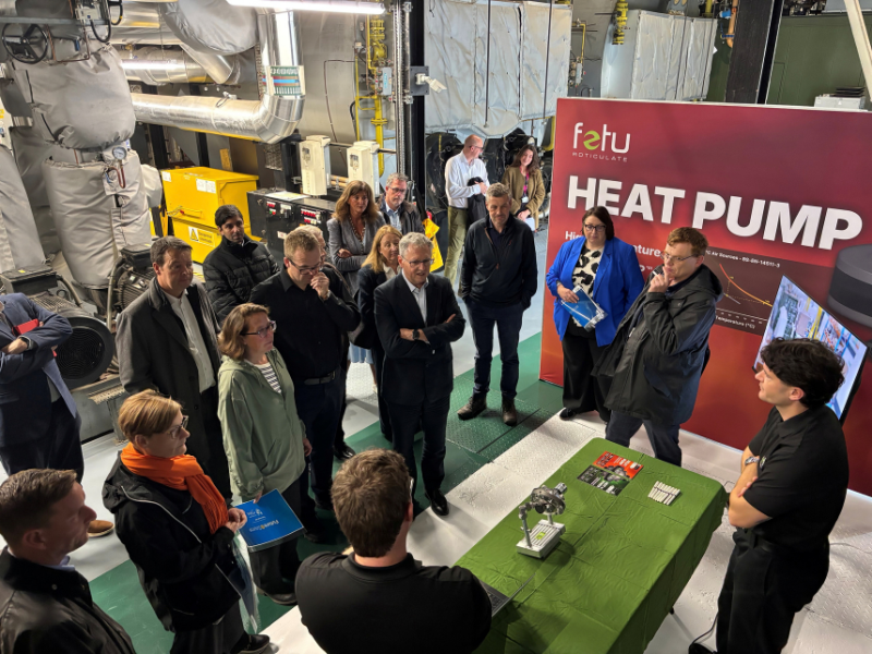 A group of people gather around a man giving a presentation.  A red wall reads "heat pump" in white letters behind them.