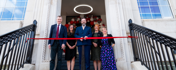 Five well-dressed people stand in a doorway at the top of a stairway preparing to cut a red ribbon.