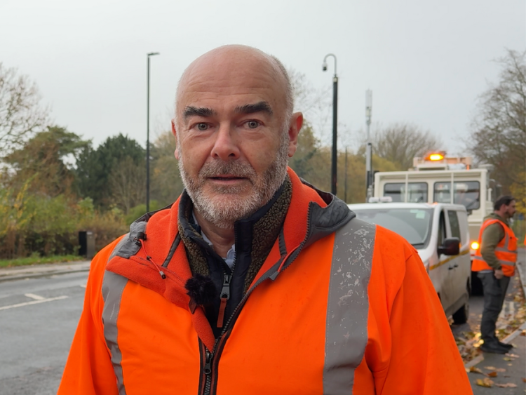 A man with a grey beard wearing an orange high-vis jacket stands in front of a road where works are taking place.