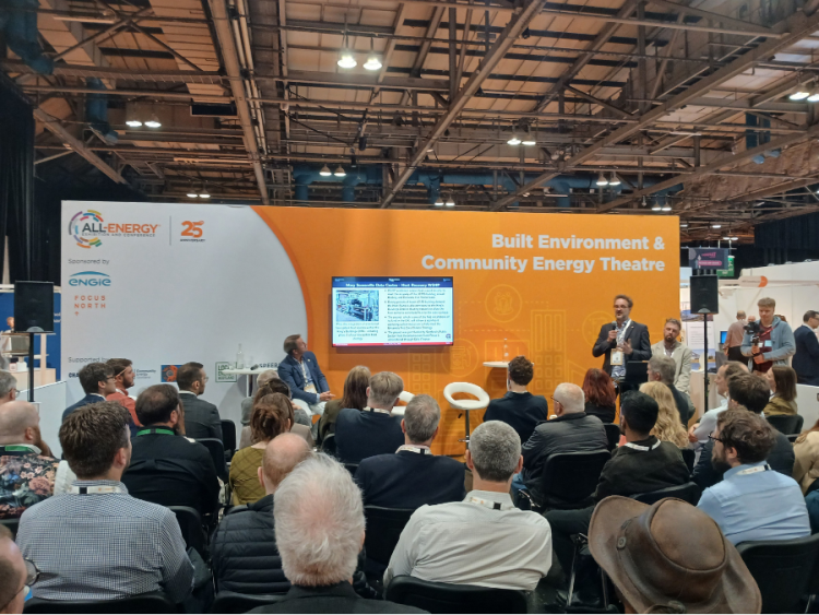 An audience in a large hall watches a man giving a presentation in front of an orange background.