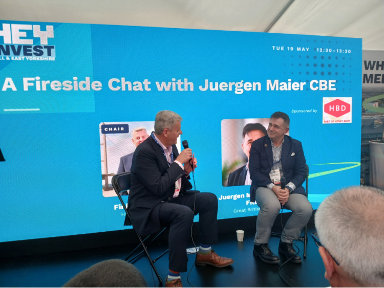 Two men sit in front of an audience with a blue background and white letters stating "a fireside chat with Jurgen Maier CBE"