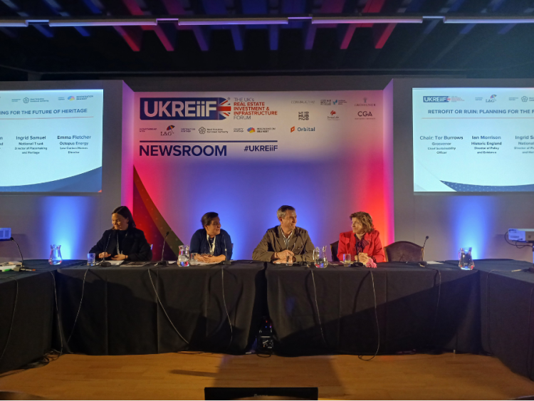 A panel of four people sit behind a table on stage with branding and screens displaying their names and titles behind them on the wall.