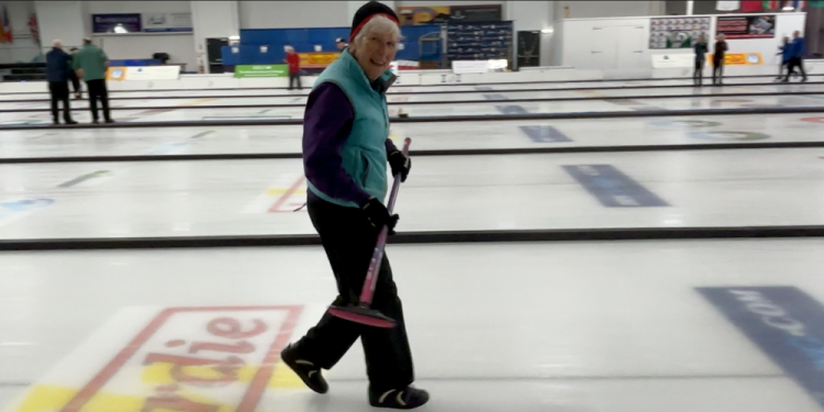A woman dressed in a tracksuit and hat, smiles while carrying her broom across the ice