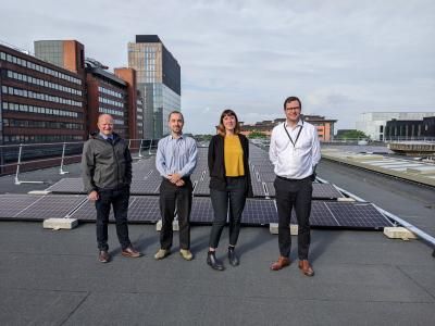 Four people on the roof of the Royal Northern College of Music standing next to roof top solar panels.